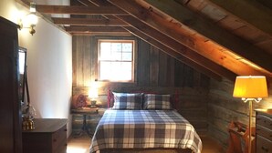 A cozy attic bedroom with a wooden floor, a checkered bedspread, a dresser, a lamp, and a window with natural light.