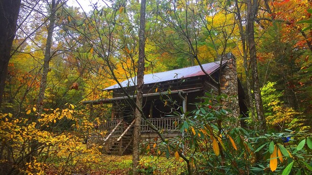 A rustic cabin with a stone foundation, wooden deck, and a metal roof, surrounded by autumn foliage.