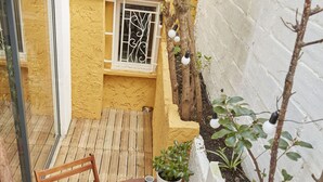Une terrasse en bois avec une table ronde et des chaises, une petite plante en pot et un mur blanc orné d’une fenêtre et d’un objet sphérique blanc.