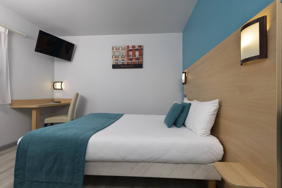 A hotel room with a bed, a desk, a chair, a TV mounted on the wall, and a picture of a building.