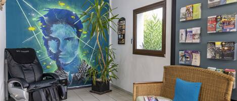 Una habitación con una silla de masajes, un sofá de mimbre con un cojín azul, un mural grande en la pared y una ventana con vista a follaje.