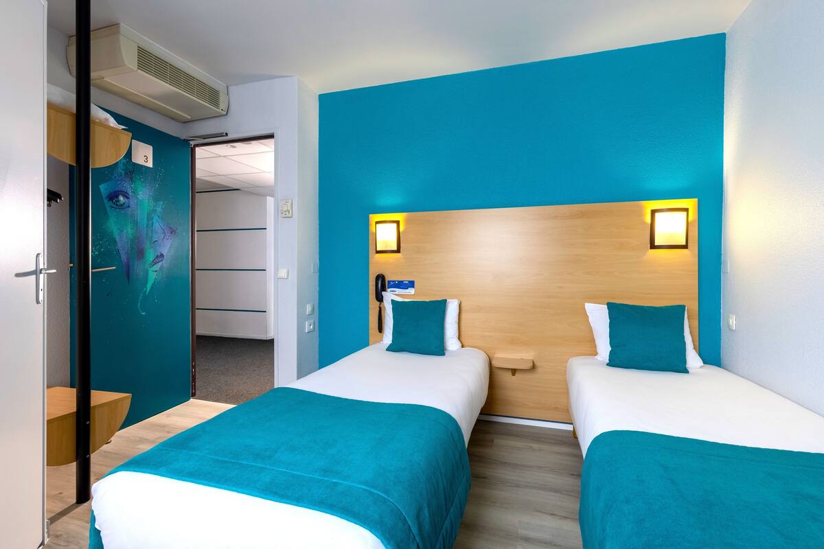 A hotel room with two beds, teal bedding, a wooden headboard, and a wall-mounted phone.
