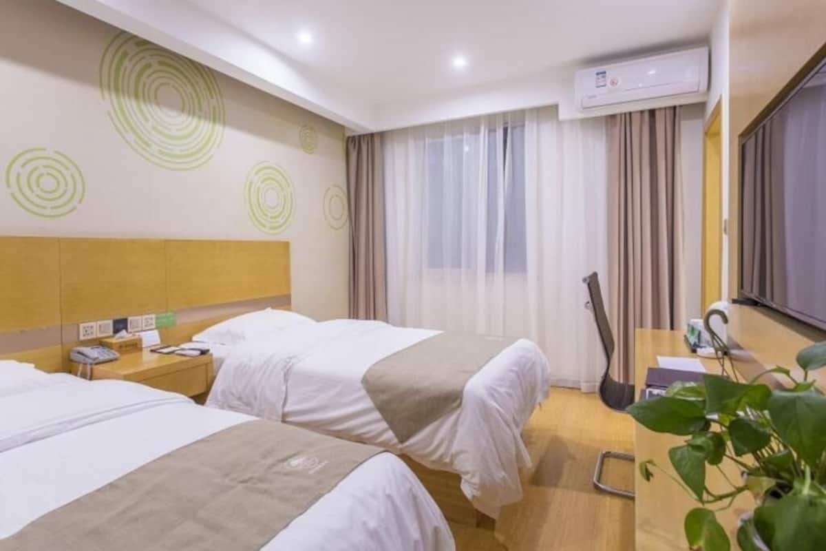 A hotel room with two beds, a desk, a chair, an air conditioning unit, and a window with curtains.