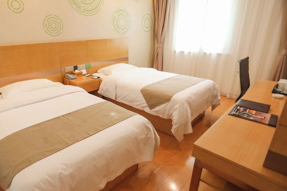 Quarto de hotel com duas camas, cabeceira de madeira, mesa de cabeceira com telefone e lenços, e escrivaninha de madeira com uma revista.