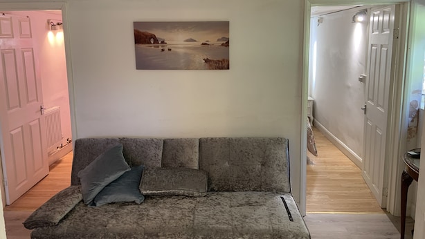 A living room with a grey sofa, a painting of a coastal scene, and a hallway leading to a door. - 2bed Room Small Annex Furnished in High Wycombe (High Wycombe)