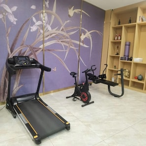 Uma academia em casa com esteira e bicicleta ergométrica, parede com papel de parede floral roxo e uma estante com diversos objetos.