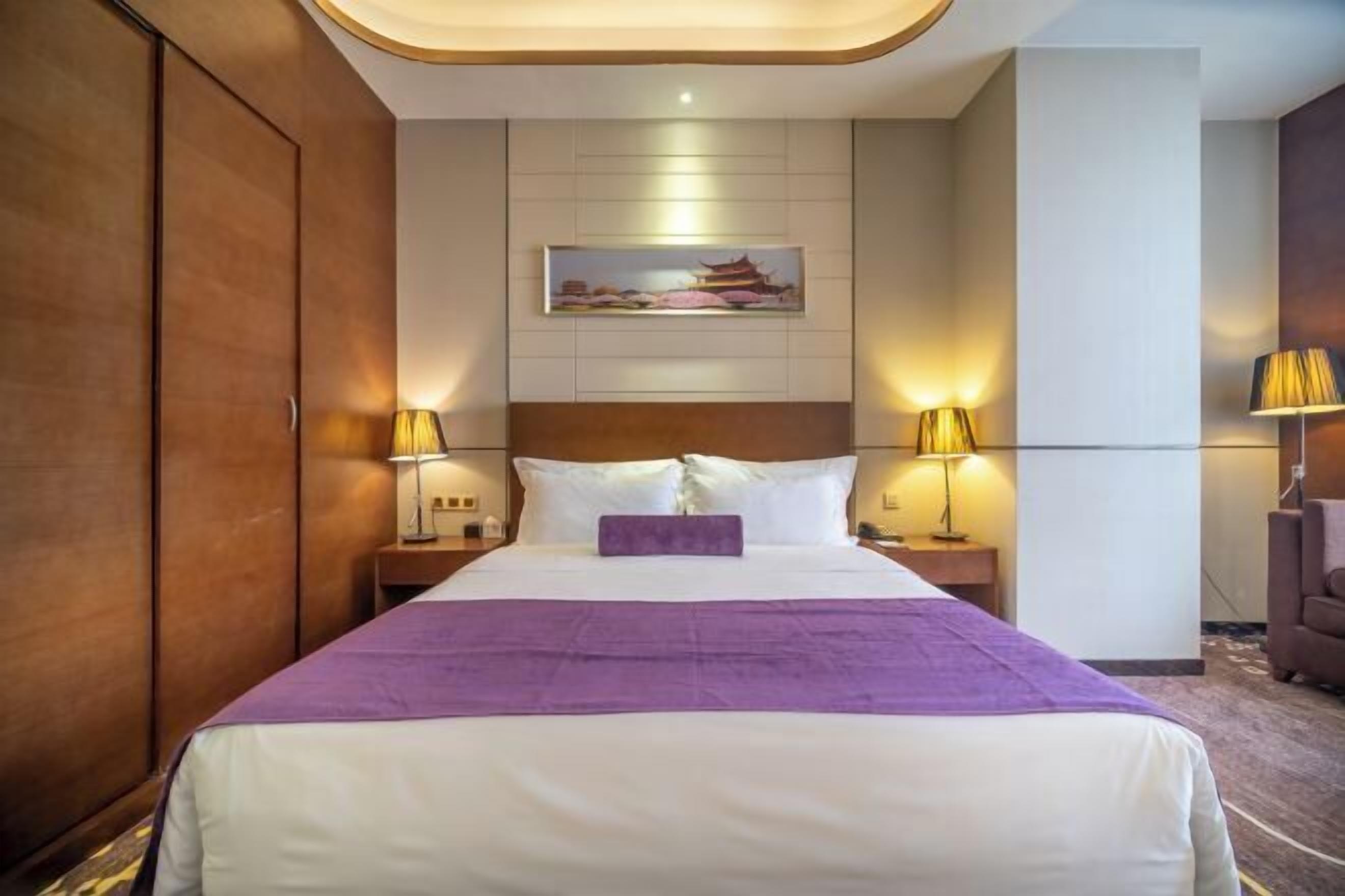 A hotel room with a large bed, two bedside tables with lamps, a headboard, a wall-mounted painting, and a wardrobe.