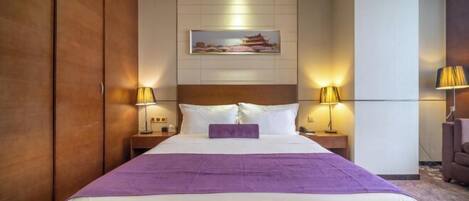 A hotel room with a large bed, two bedside tables with lamps, a headboard, a wall-mounted painting, and a wardrobe.