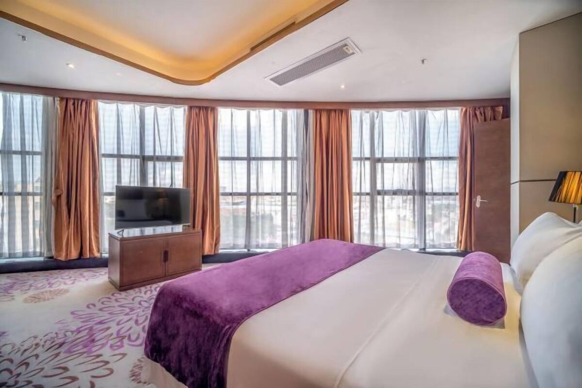 A hotel room with a large bed, a television, a wardrobe, and large windows with curtains.
