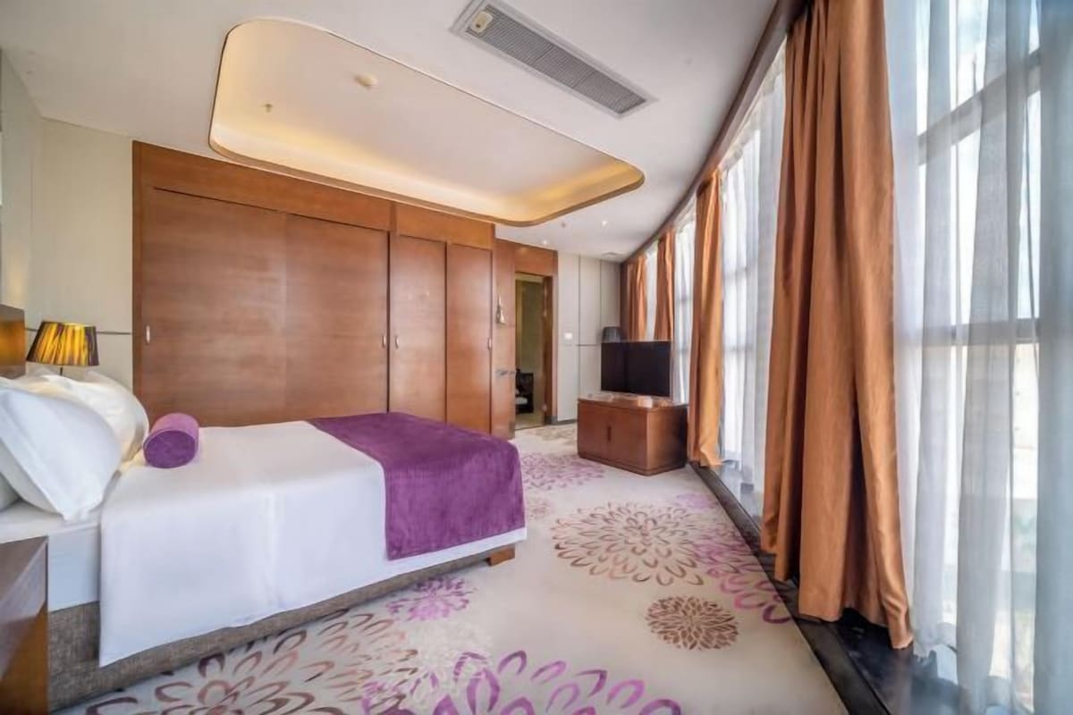A hotel room with a bed, a TV, a wardrobe, and a large window with curtains.