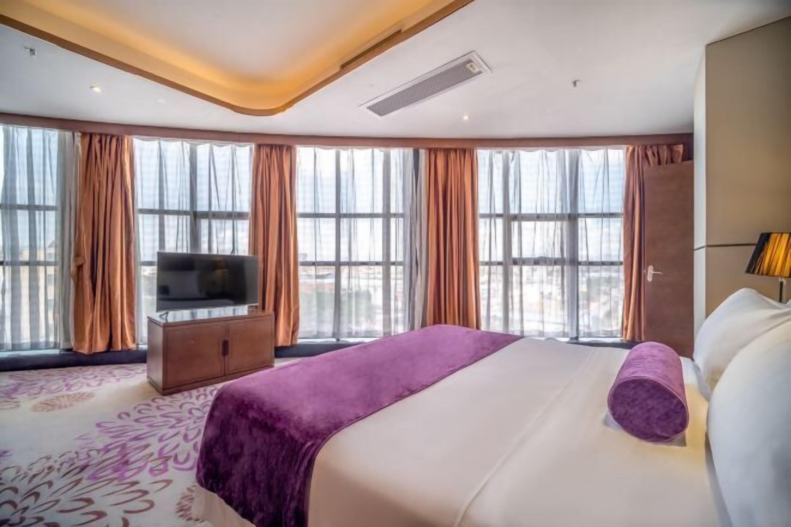 A hotel room with a large bed, a TV, and a view of the city through large windows.