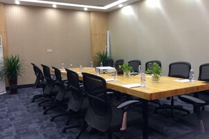 Un salón de conferencias con una larga mesa, sillas negras, plantas en macetas y botellas de agua.