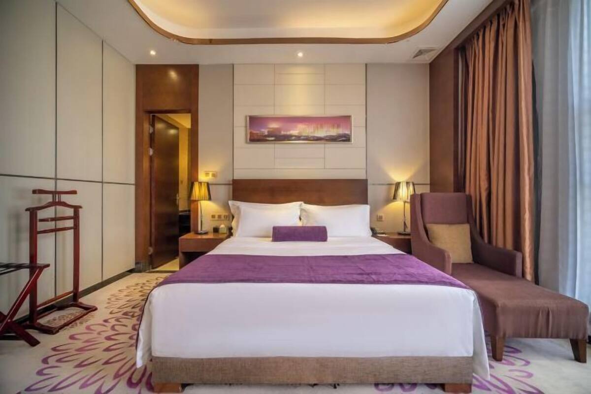 A hotel room with a large bed, a headboard, two bedside tables with lamps, a chair, and a painting on the wall.