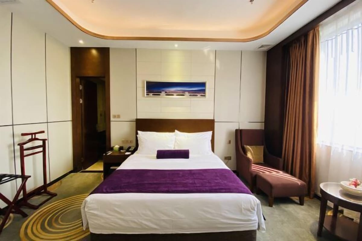 A hotel room with a large bed, a nightstand, a chair, a small table, and a window with curtains.