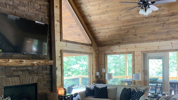 A spacious living area with a stone fireplace, a large flat-screen TV mounted on a brick wall, a wooden ceiling, and a ceiling fan. - BUDDHA BLUE RIDGE TREES is a BRAND NEW custom 4/4 Close to town Find your ZEN... (Blue Ridge)