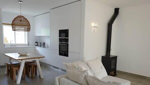 A modern kitchen with white cabinetry, a built-in oven, and a black stove. There is a dining area with a wooden table and chairs, and a white sofa in the living area. - Jardins Vale de Parra (Albufeira)