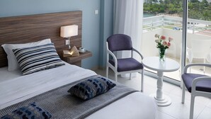 Habitación de hotel con cama, mesita de noche con lámpara, una silla, una pequeña mesa con un florero y un balcón con vistas al mar y la vegetación.