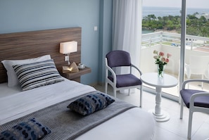 Une chambre d’hôtel comprenant un lit, une table de chevet avec une lampe, une chaise, une petite table avec un vase de fleurs et un balcon offrant une vue sur la mer et la verdure.
