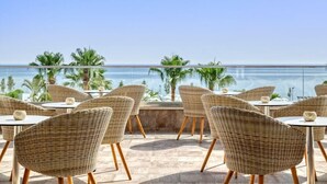 A terrace with wicker chairs and tables, overlooking the sea with palm trees in the background. - Atlantica Sea Breeze - Adults Only (Paralimni)