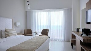 Habitación de hotel con una cama grande, dos sillones, una mesita, un escritorio con televisor y vistas al mar a través de cortinas translúcidas.