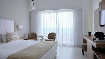 A hotel room with a large bed, two armchairs, a small table, a desk with a TV, and a view of the sea through sheer curtains.