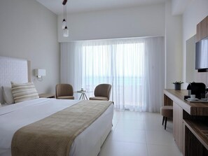Une chambre d’hôtel avec un grand lit, deux fauteuils, une petite table, un bureau avec une télévision et une vue sur la mer à travers de fines rideaux.