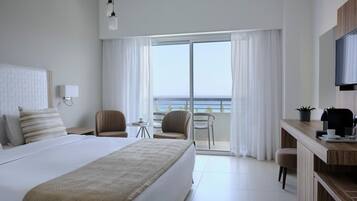 A modern hotel room with a large bed, a desk with a coffee maker, and a view of the sea through the balcony.
