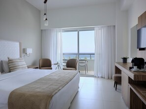 Une chambre d’hôtel moderne dotée d’un grand lit, d’un bureau avec une machine à café et offrant une vue sur la mer depuis le balcon.