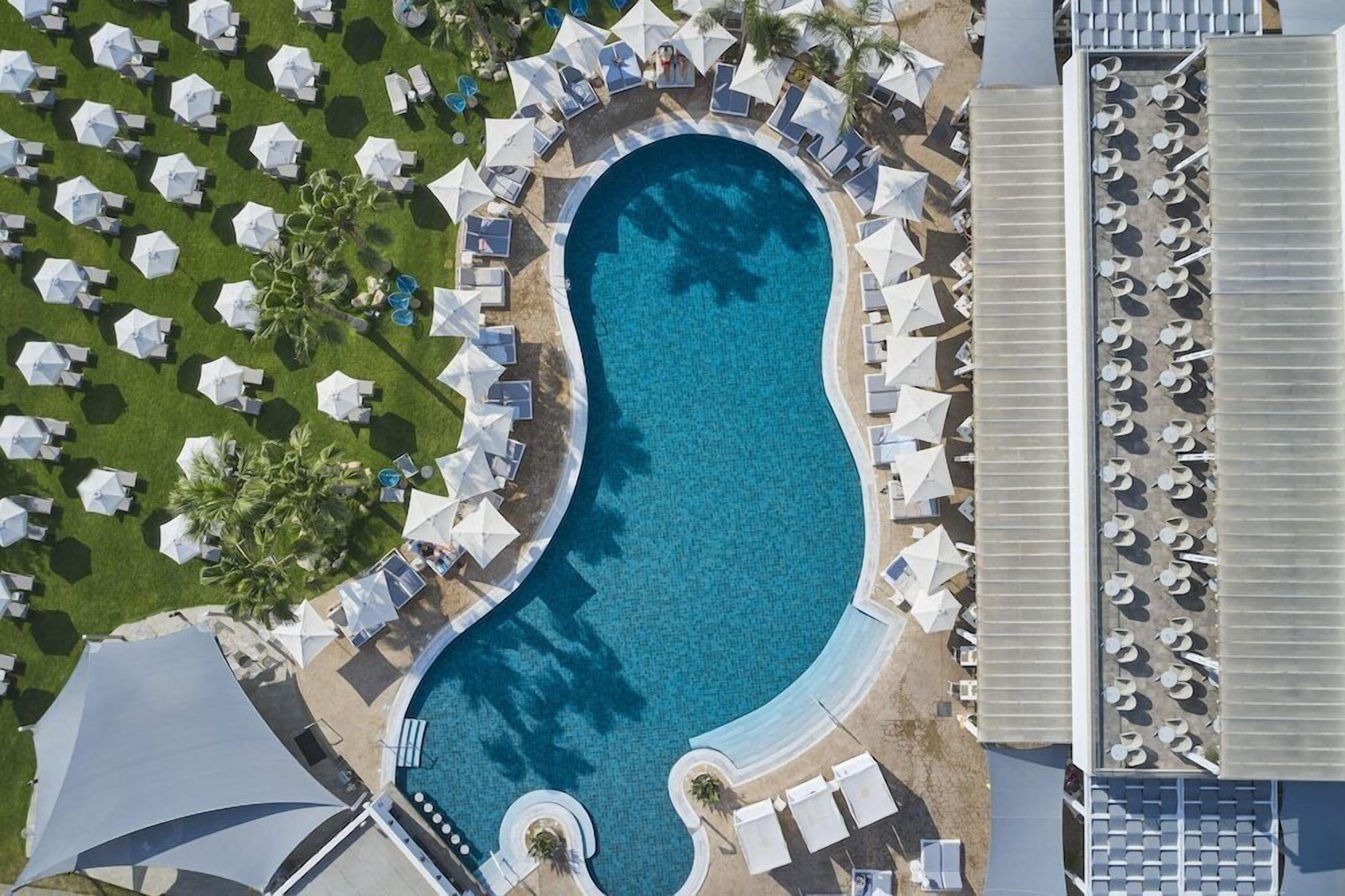 An aerial view of a resort with a large swimming pool surrounded by sun loungers and umbrellas, a covered seating area, and a section with additional sun loungers.