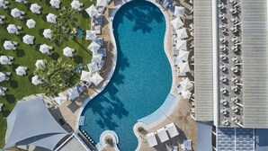 Vista aérea de un resort con una gran piscina rodeada de sillones de baño y sombrillas, un área de estar techada y una sección con más sillones de baño.