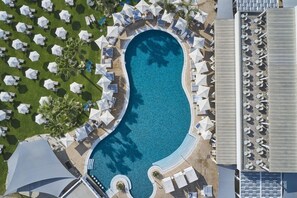 Vue aérienne d’un complexe hôtelier doté d’une grande piscine entourée de transats et de parasols, d’un espace de détente couvert et d’une zone avec des transats supplémentaires.
