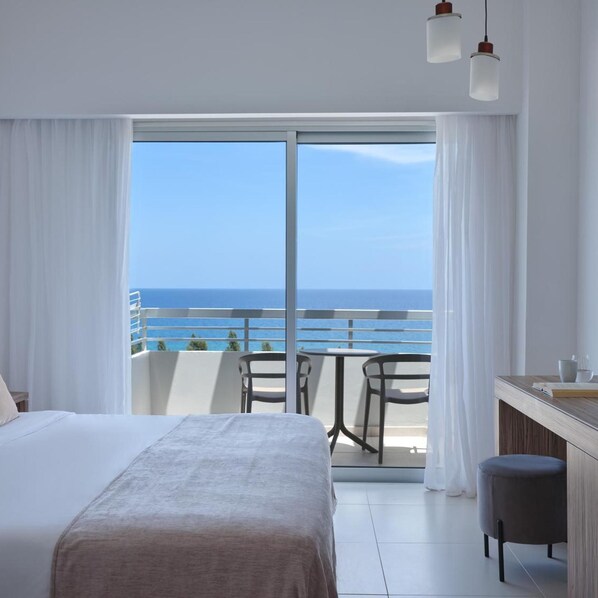 A hotel room with a bed, a small table, a chair, a bench, and a view of the ocean.