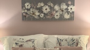 Una cama con ropa blanca, dos almohadas con forma de corazón que tienen la palabra 'happy' y un cuadro floral sobre la cama.