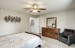 Un dormitorio con cama, cómoda, tocador con espejo, ventilador de techo y puerta que da a otra habitación.