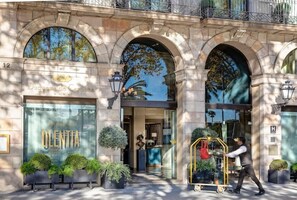 Entrada de un hotel con un amplio ventanal de vidrio, un letrero que dice 'OLENTIA' y un hombre empujando un carrito.
