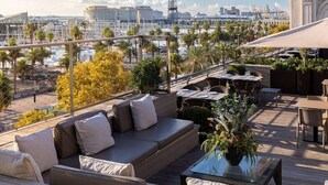 A rooftop terrace with a sofa, dining table, and chairs, overlooking a marina with boats and palm trees. - Duquesa de Cardona (Barcelona)