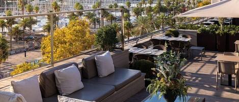 A rooftop terrace with a sofa, dining table, and chairs, overlooking a marina with boats and palm trees.