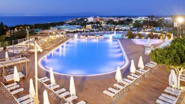 A large outdoor swimming pool with lounge chairs and umbrellas, surrounded by buildings and a view of the sea. - Kipriotis Panorama and Suites (Kos)