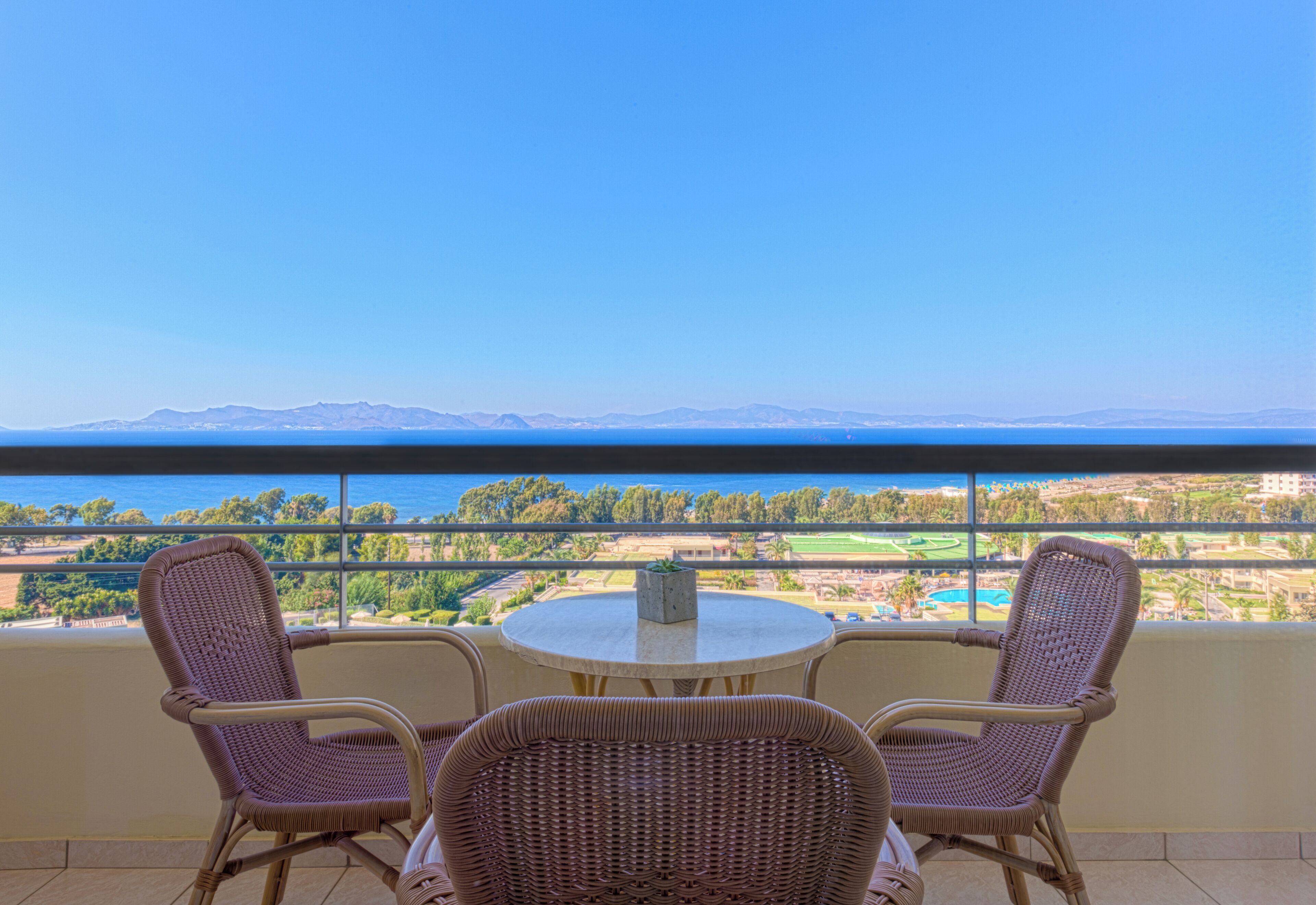 Ein Balkon mit Korbstühlen und einem Tisch, mit Blick auf eine Küstenlandschaft mit Bergen und viel Grün.