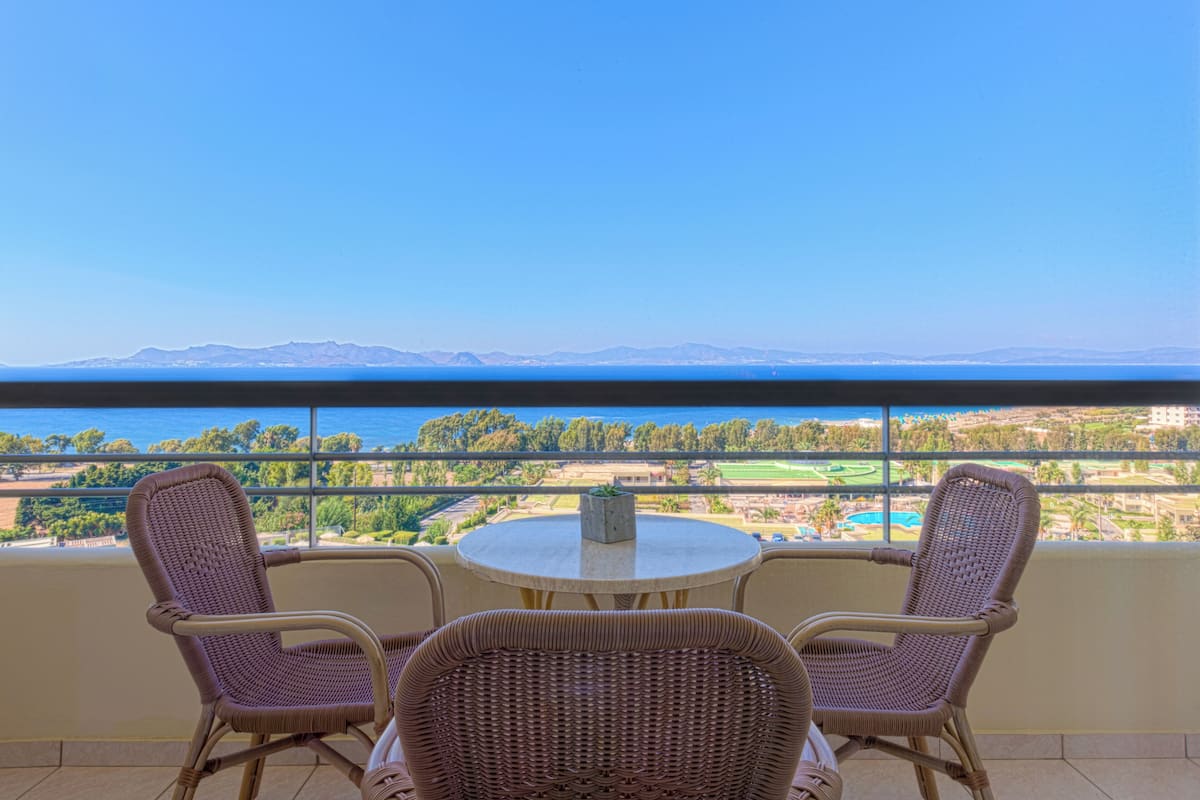 A balcony with wicker chairs and a table, overlooking a coastal landscape with mountains and greenery.