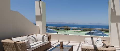 A balcony with wicker furniture, a view of the sea, and a tennis court.