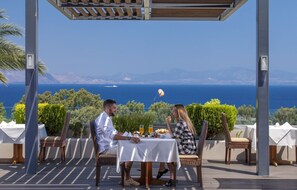 Un couple est assis à une table, devant laquelle s’étend un paysage offrant une vue sur la mer et les montagnes.