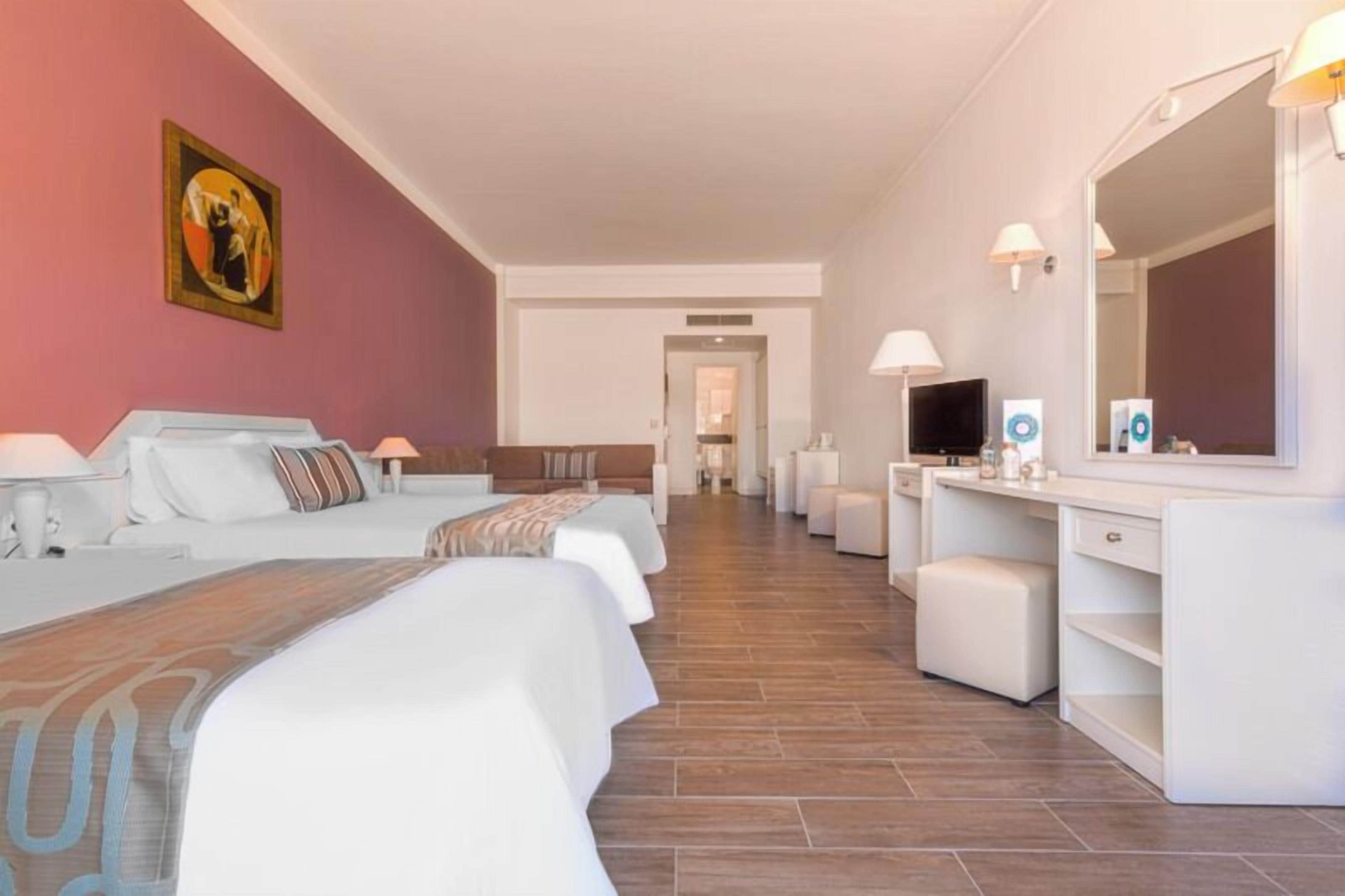 Ein Hotelzimmer mit einem langen Bett, einem Schreibtisch mit Spiegel, einem Fernseher und einem im Hintergrund sichtbaren Badezimmer.