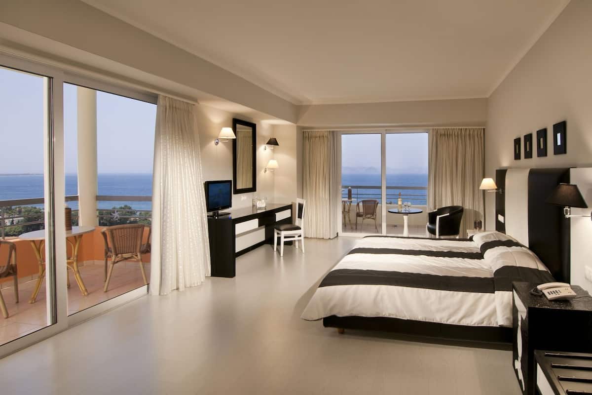 A modern hotel room with a large bed, a desk, a TV, and a balcony with a view of the sea.