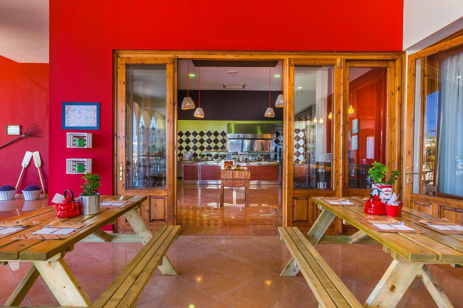 A restaurant with wooden tables and benches, a red wall, and a kitchen in the background.