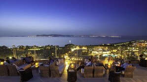 A rooftop terrace with wicker furniture, a view of the sea, and city lights in the background. - Kipriotis Panorama and Suites (Kos)