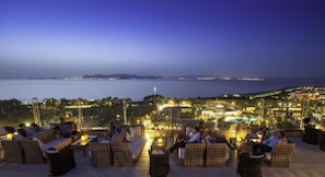 A rooftop terrace with wicker furniture, a view of the sea, and city lights in the background.