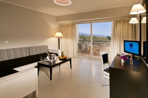 Habitación de hotel con sofá, mesa de comedor con frutero, televisión y balcón con vistas.