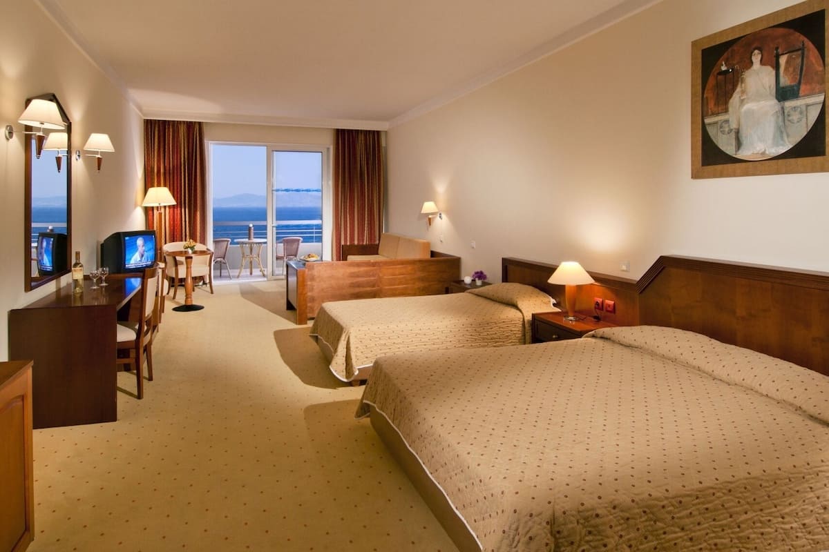 A hotel room with two beds, a desk with a television, a dining area with chairs, and a view of the sea.