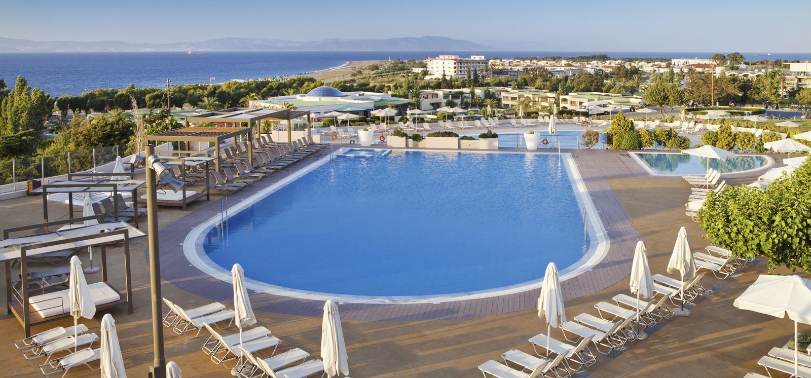 A large pool with sun loungers and umbrellas, surrounded by buildings and a view of the sea.